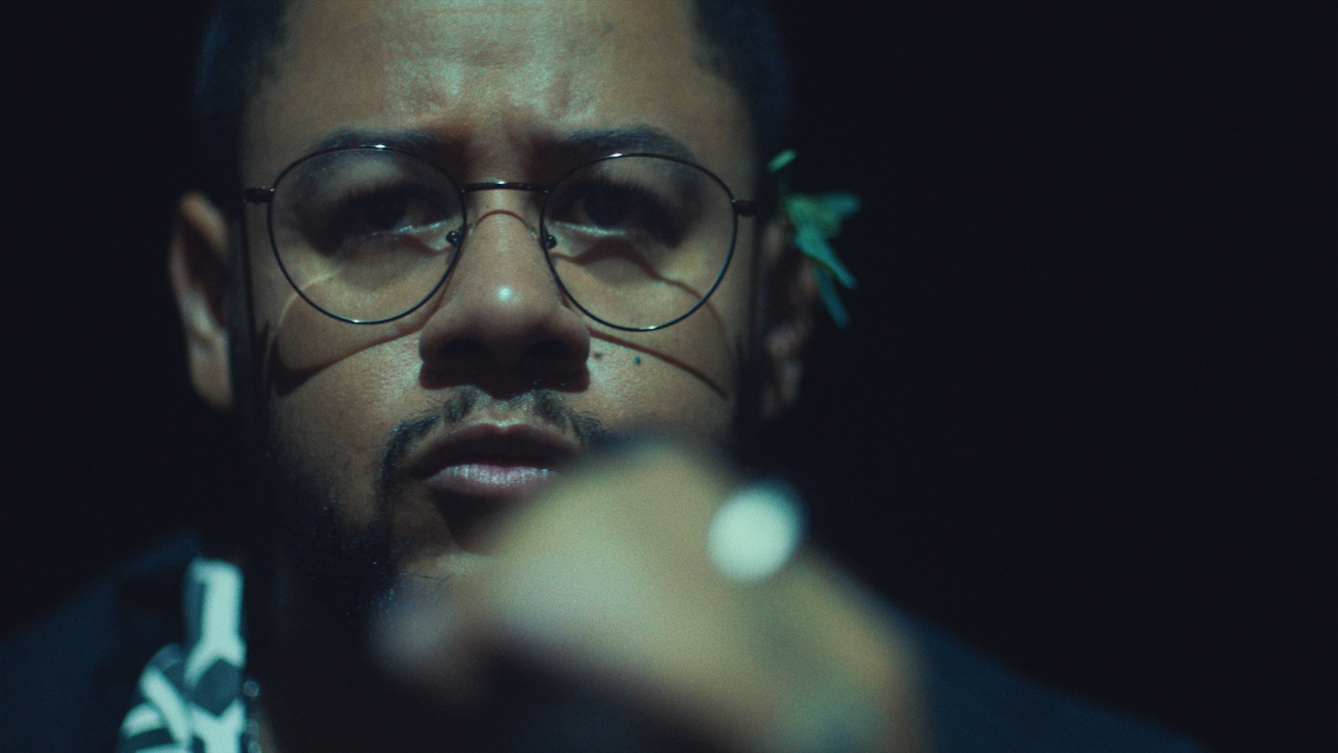 Emicida portrait — moody close-up frame from the AmarELO Silêncio campaign, wearing glasses, dark teal lighting