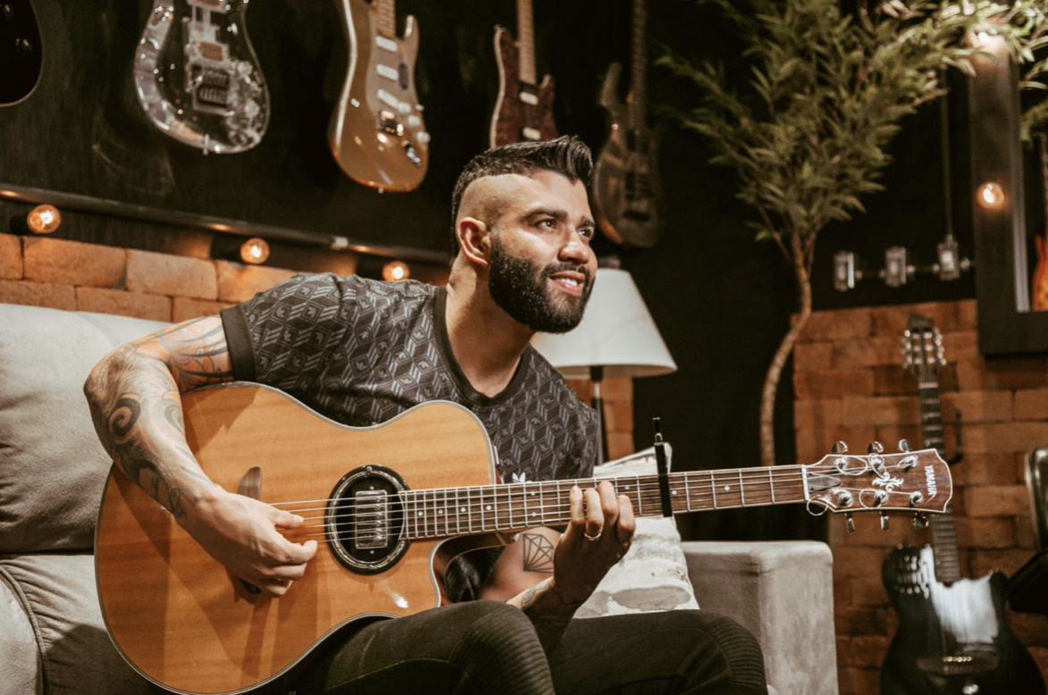 Gusttavo Lima playing acoustic guitar at a Deezer Sessions studio with a wall of guitars behind him
